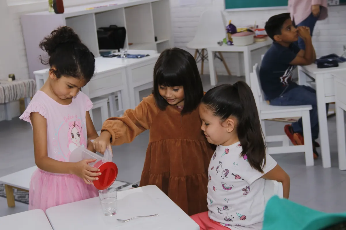 Niñas sirviendo agua con una pequeña jarra de plástico en un vaso de cristal.