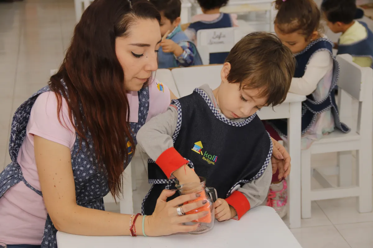 Guía Montessori sosteniendo una jarra de vidrio a un niño que la seca con un trapo.