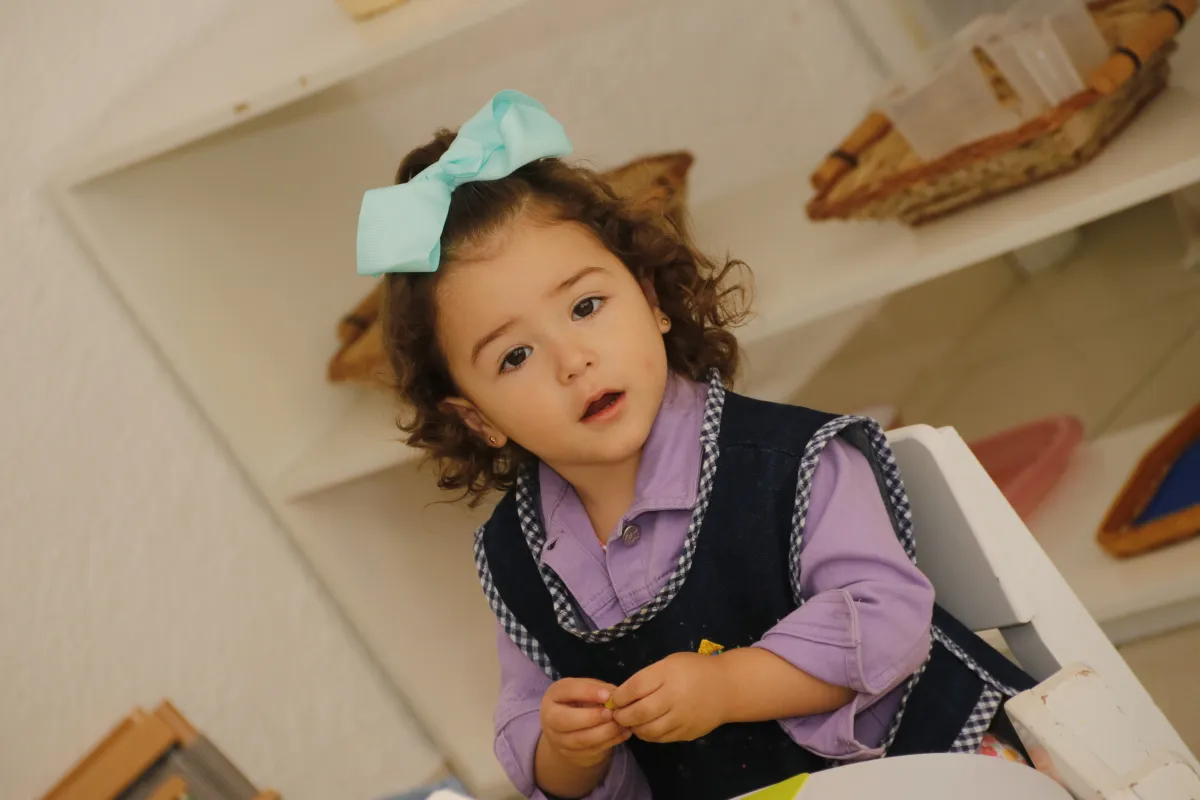 Pequeña niña con un moño turquesa en el cabello sentada frente a una mesita.